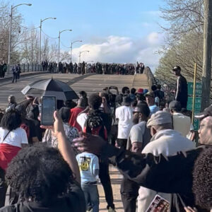 A crowd watching another crowd marching across a bridge.