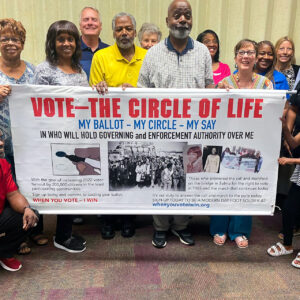 A smiling group holds a banner with the title, Vote: The Circle of Life.