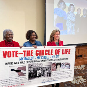 Three women hold a banner titled, Vote—The Circle of Life.