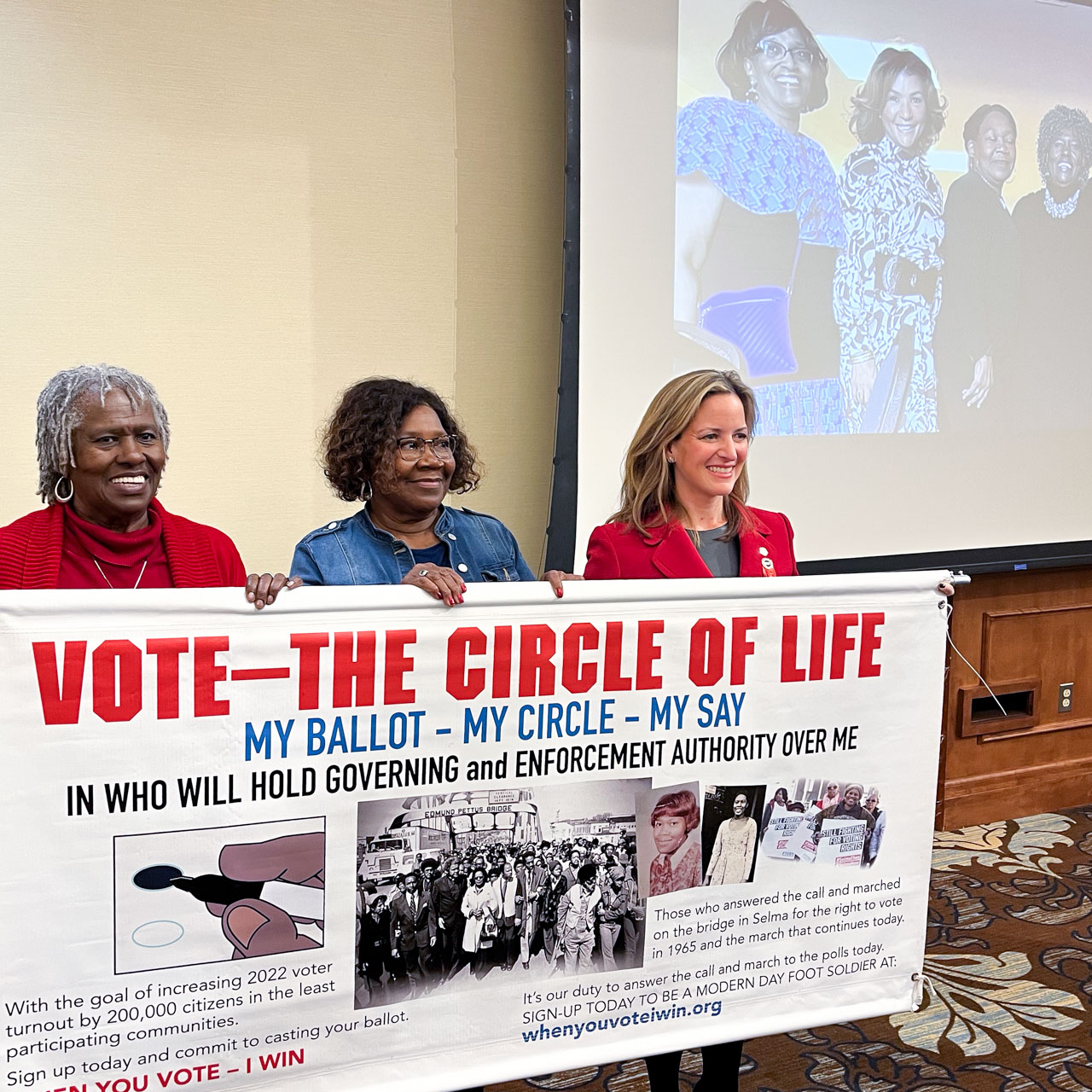 Three women hold a banner titled, Vote—The Circle of Life.
