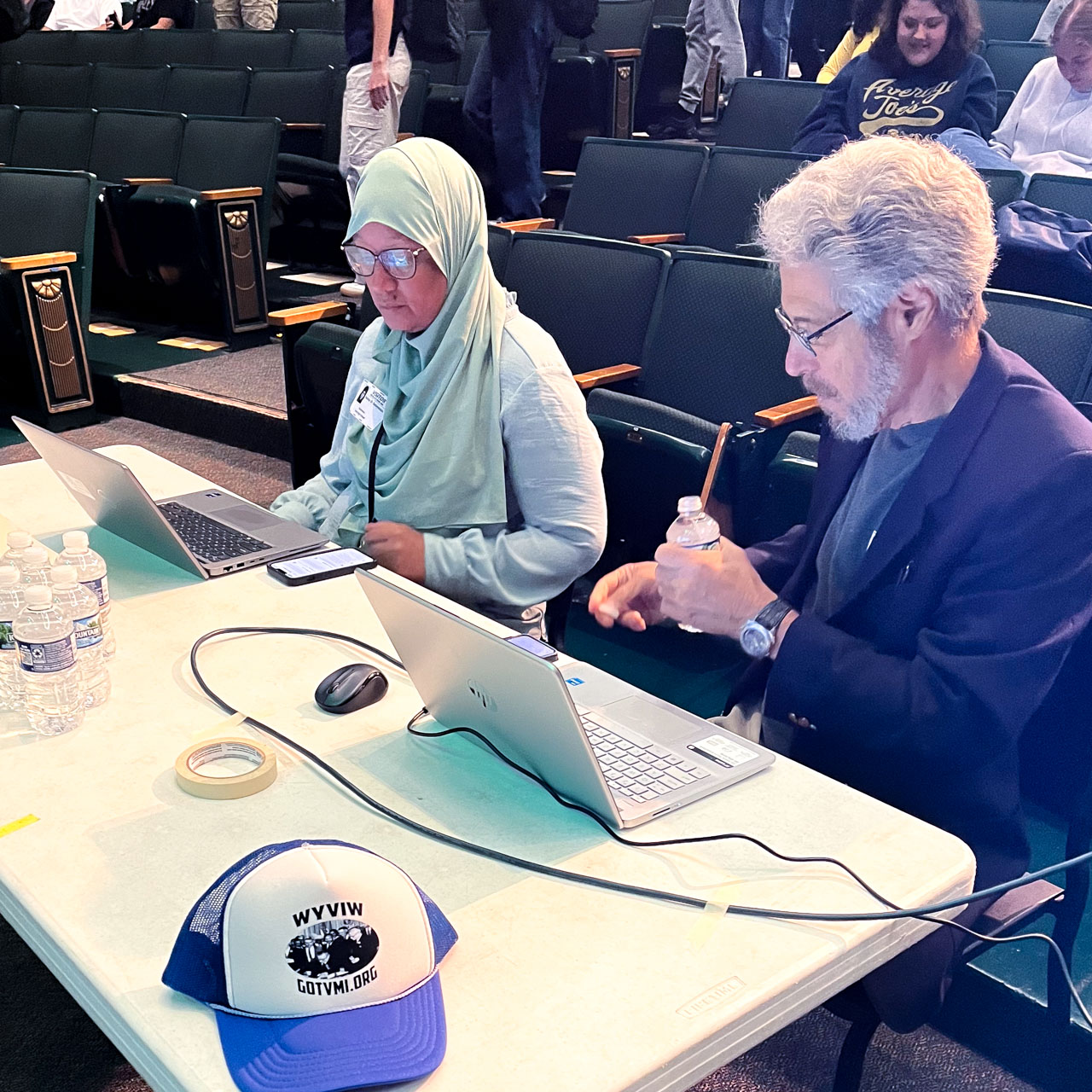 Two people seated at a table in front of laptops prepare for a presentation in an audiorium.