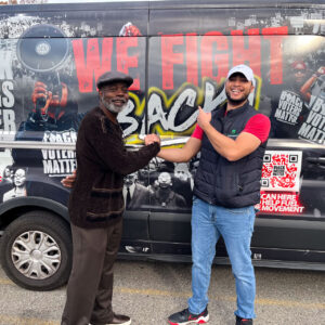 Two men smile and grasp hands in front of a van printed with voting slogans.