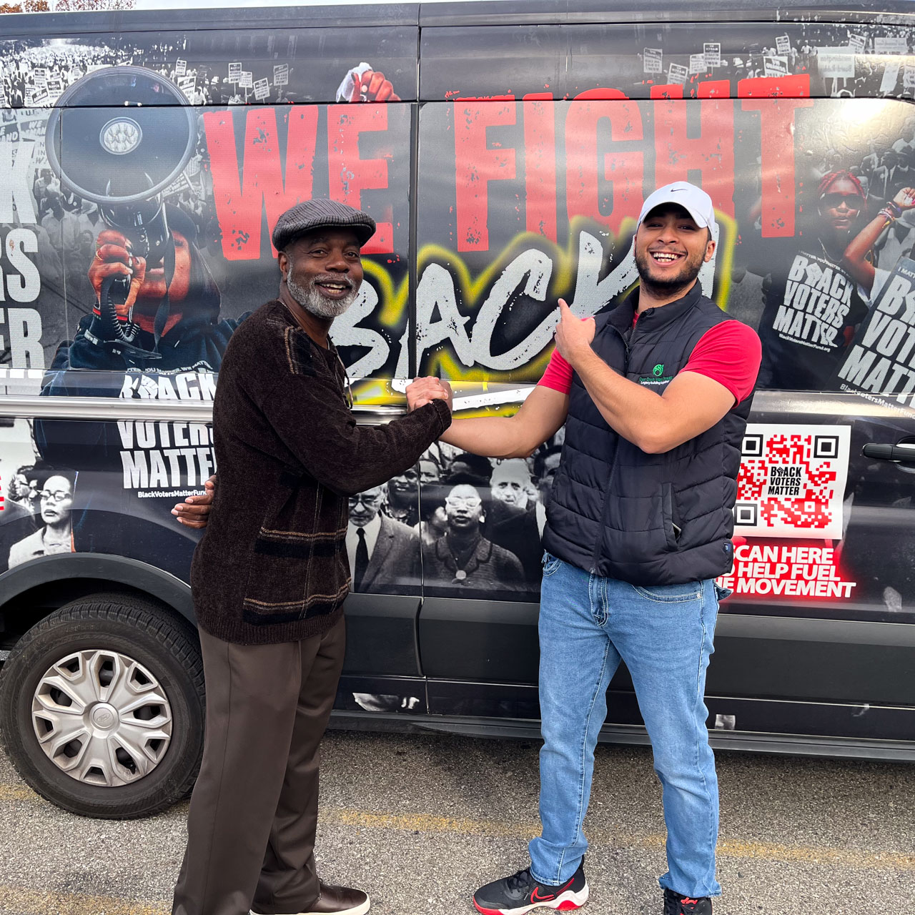 Two men smile and grasp hands in front of a van printed with voting slogans.