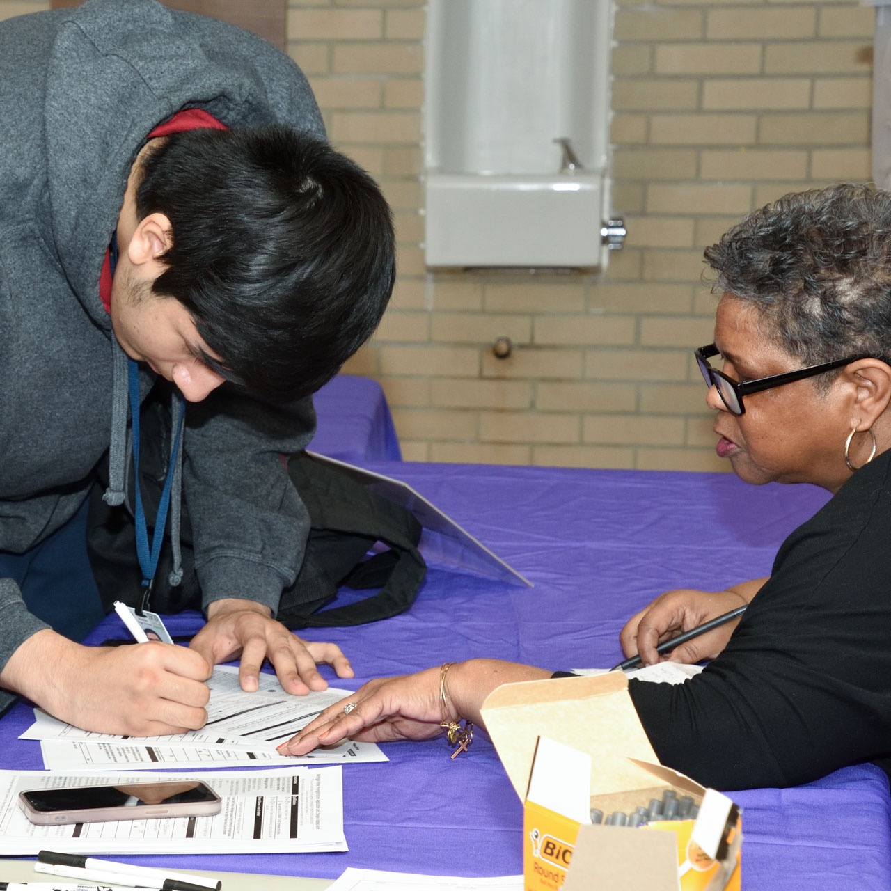 A young man bends over a table signing papers at the direction of an older woman.