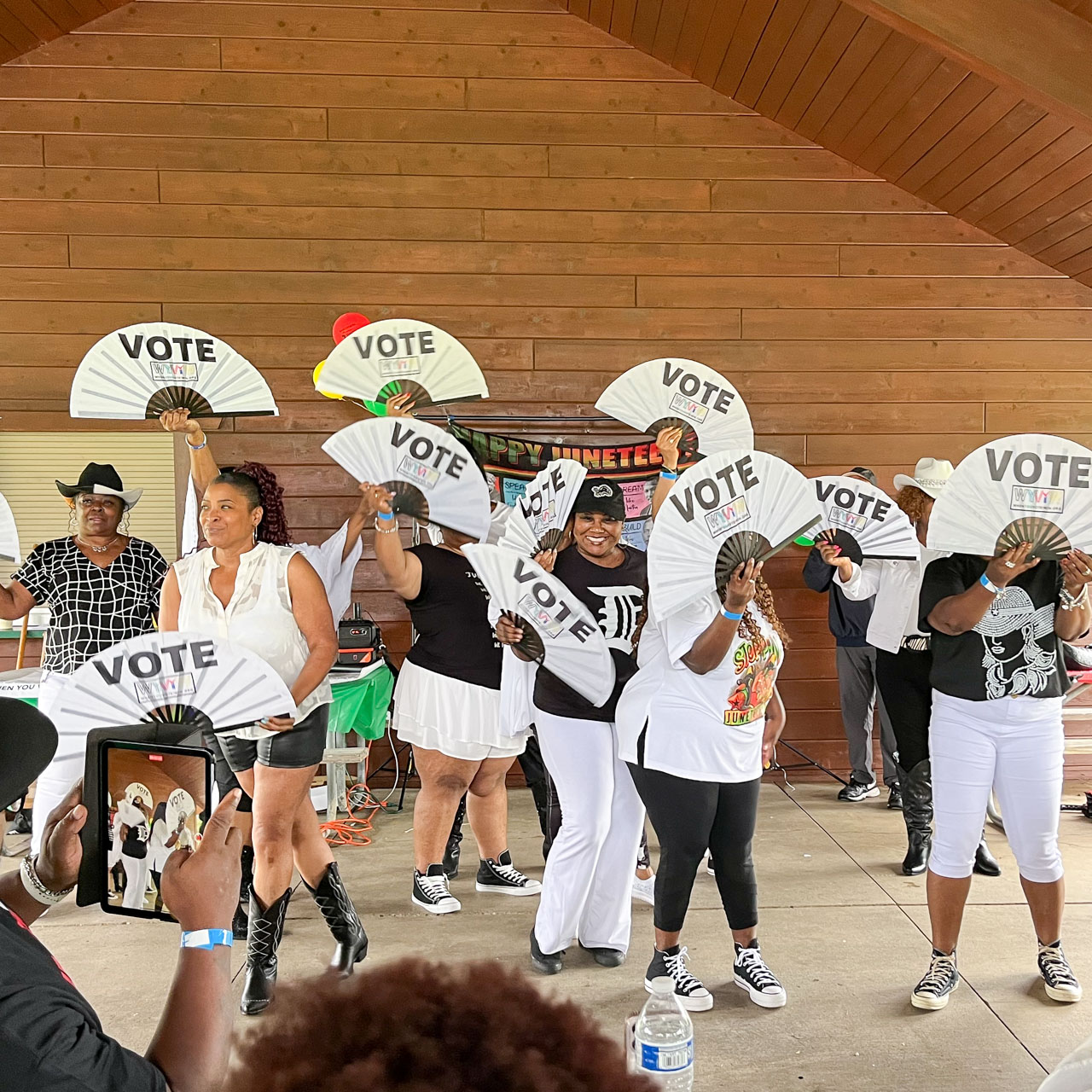 A group of people in a shelter waving fans printed with the word, vote.